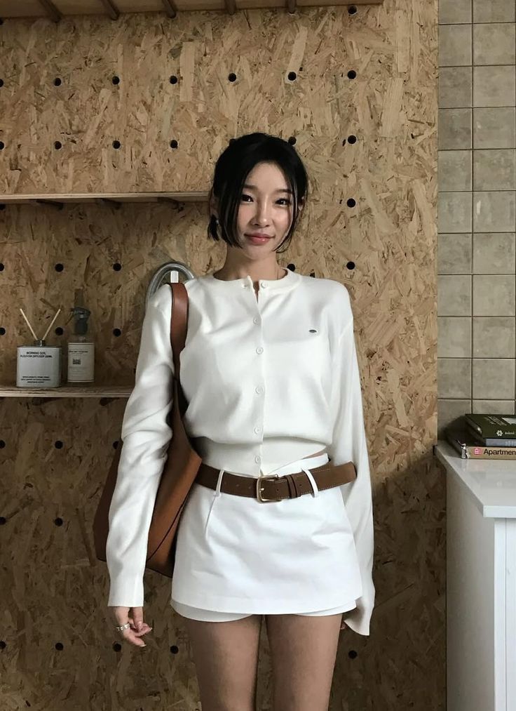 Woman in a white cardigan and skirt with a brown belt and bag indoors