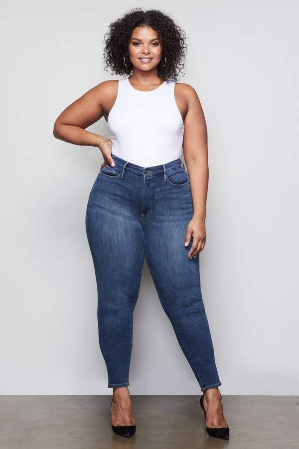 Woman with curly hair wearing a white tank top and blue jeans, smiling at the camera