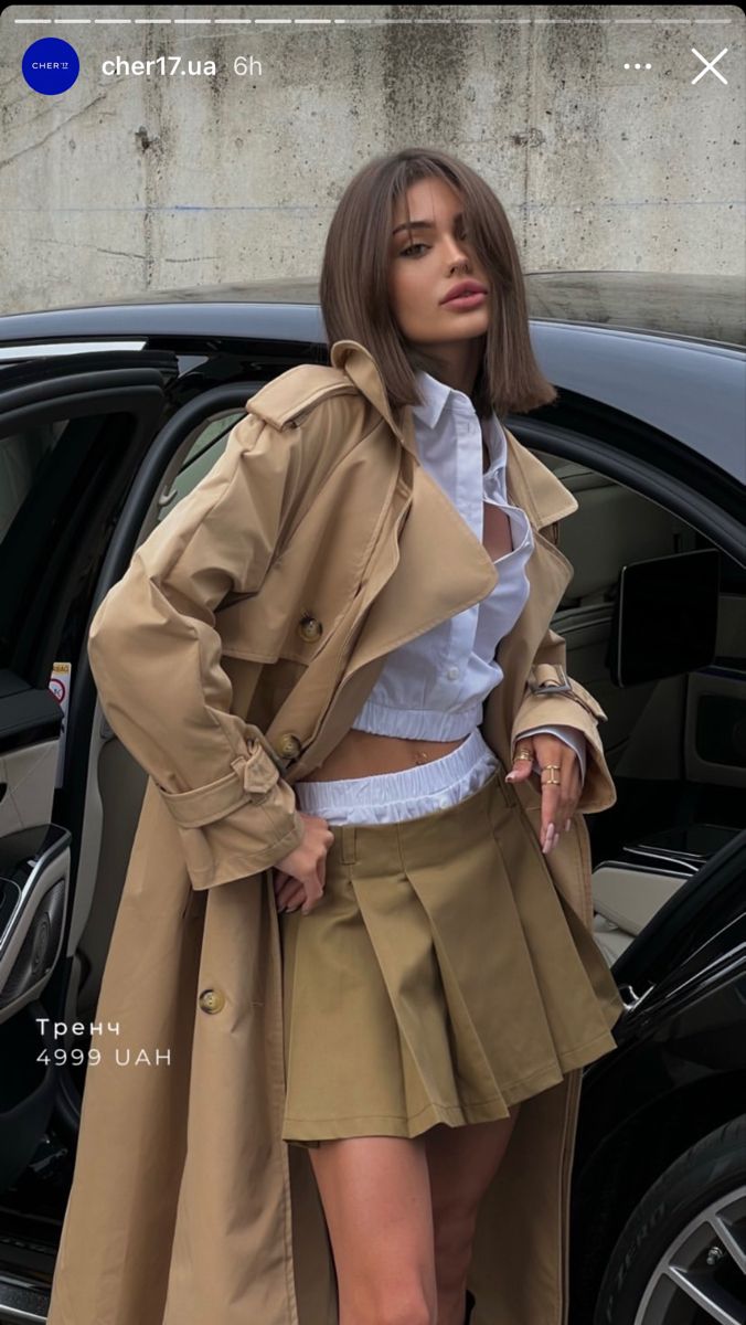 Woman in a trench coat, crop top, and pleated skirt next to a car