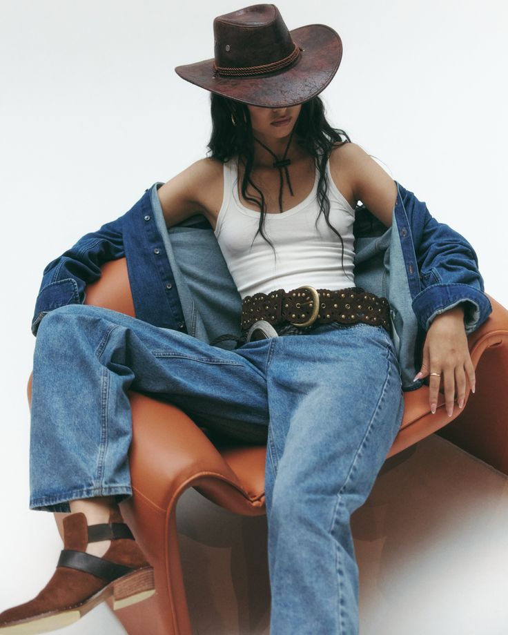 Person in a cowboy hat, denim jacket, and jeans sitting on a brown chair