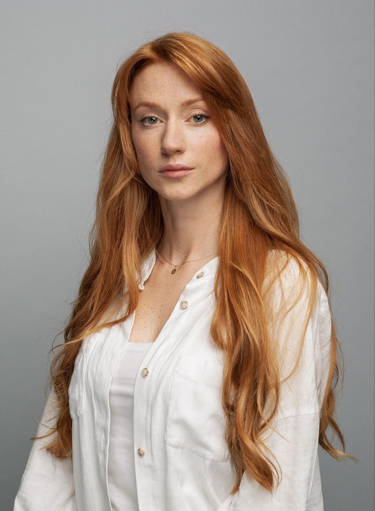 Woman with long red hair and freckles wearing a white shirt against a gray background
