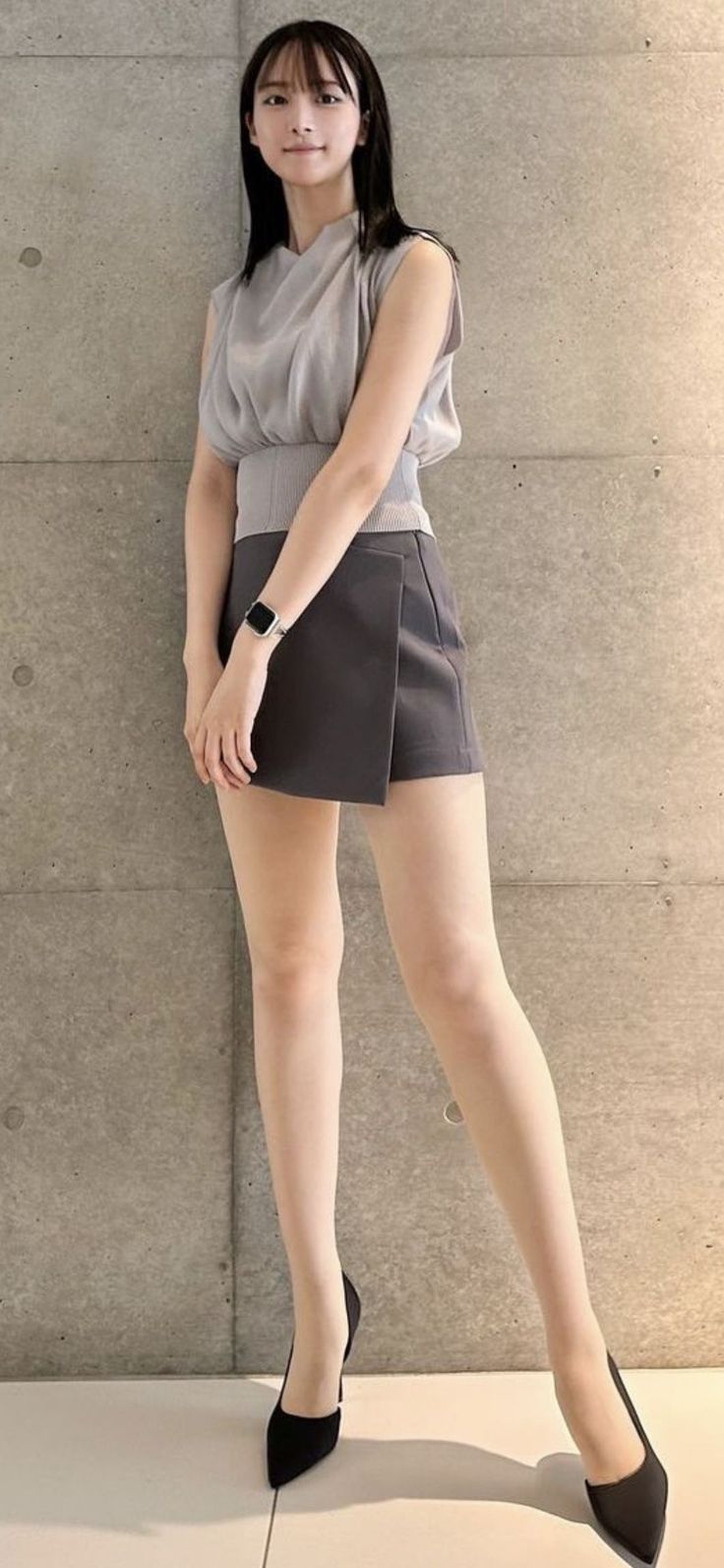 Woman with black hair wearing a short skirt and high heels against a concrete wall