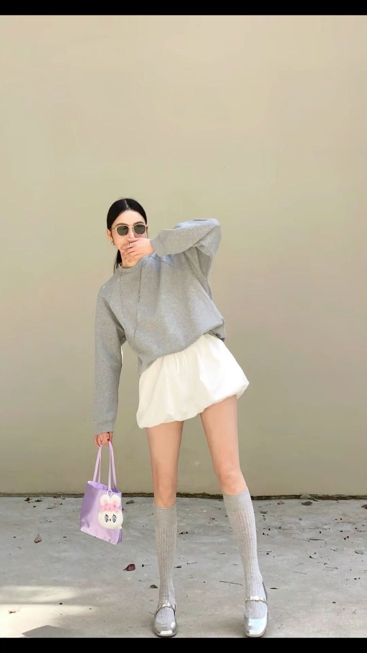 Woman in a grey sweatshirt and white skirt holding a purple bag outdoors