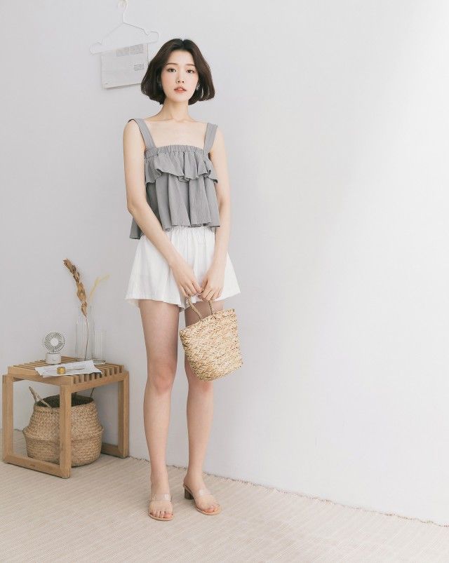 Woman with short hair wearing a ruffle top and white shorts, holding a straw bag