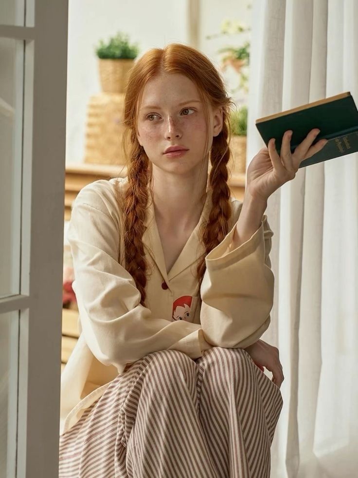 Woman with red braided hair and freckles wearing pajamas, holding a book indoors