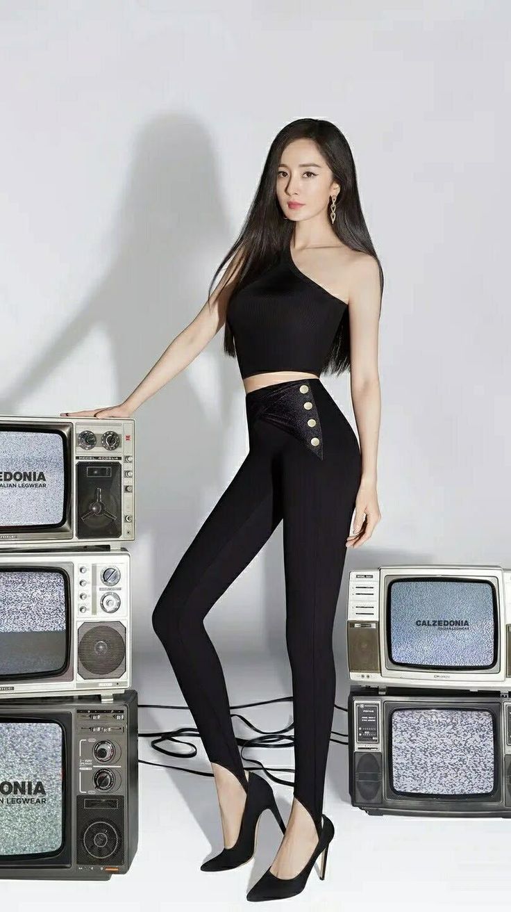 Woman in a black one-shoulder top and leggings posing with retro TVs