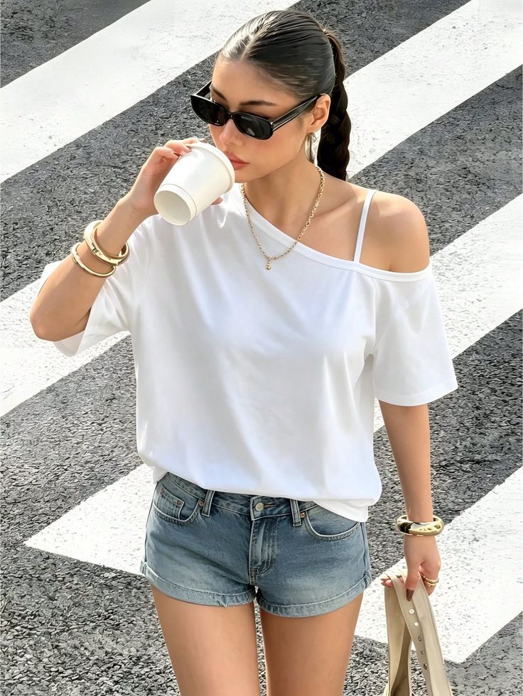 Woman in denim shorts and a white t-shirt drinking from a cup on a street