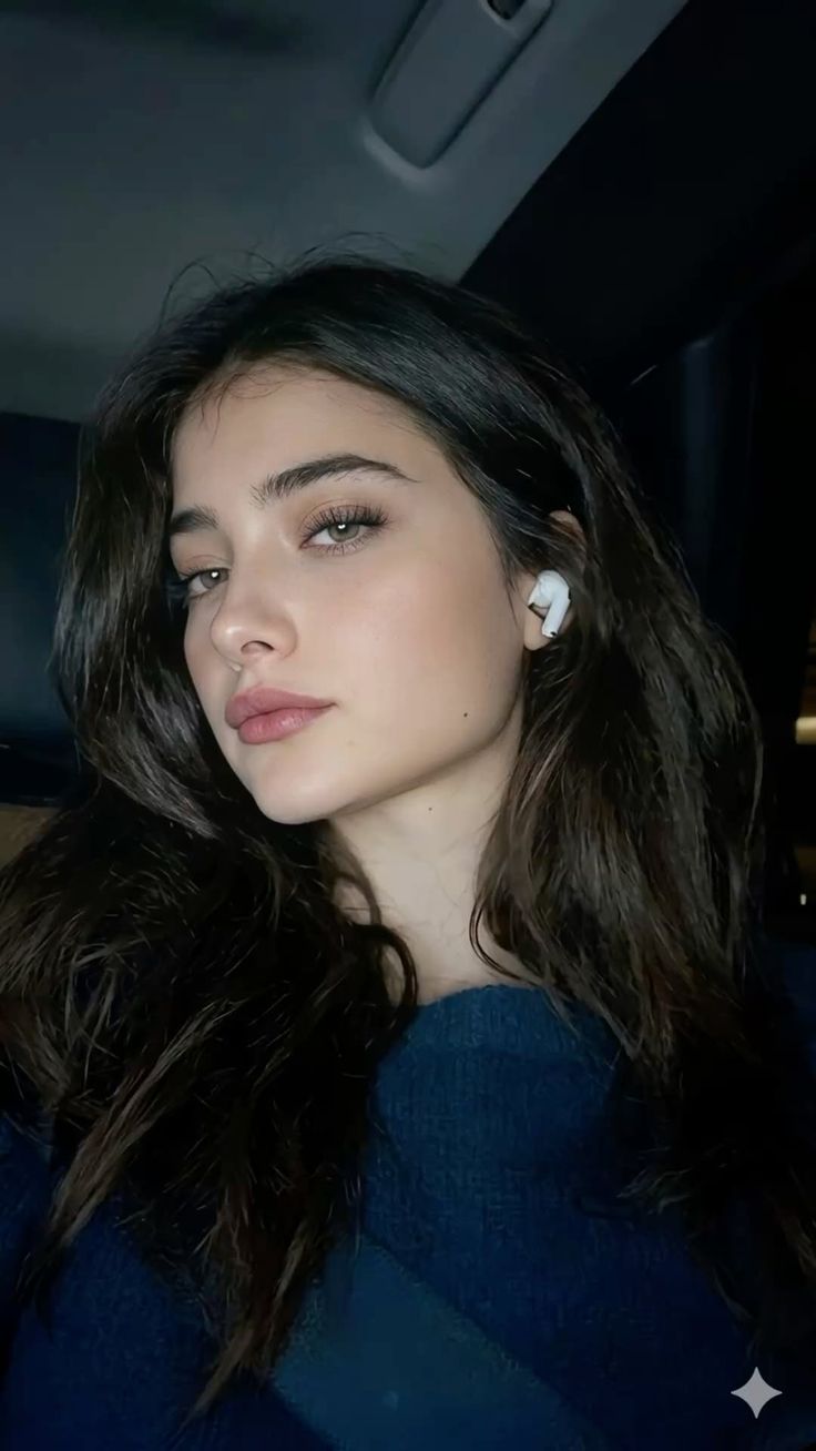 Woman with long dark hair and green eyes wearing wireless earbuds and a blue sweater