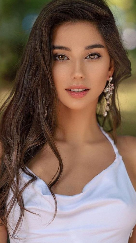 Woman with long brown hair and dangle earrings wearing a white top outdoors