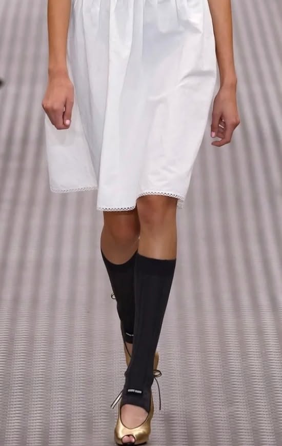 Model in a white dress, black socks, and gold peep-toe heels on a runway