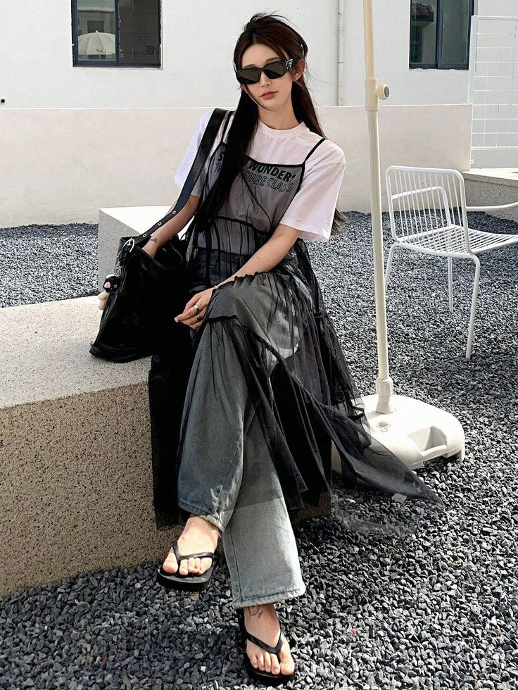 Woman in sunglasses, a white t-shirt, and a black sheer dress sitting outdoors
