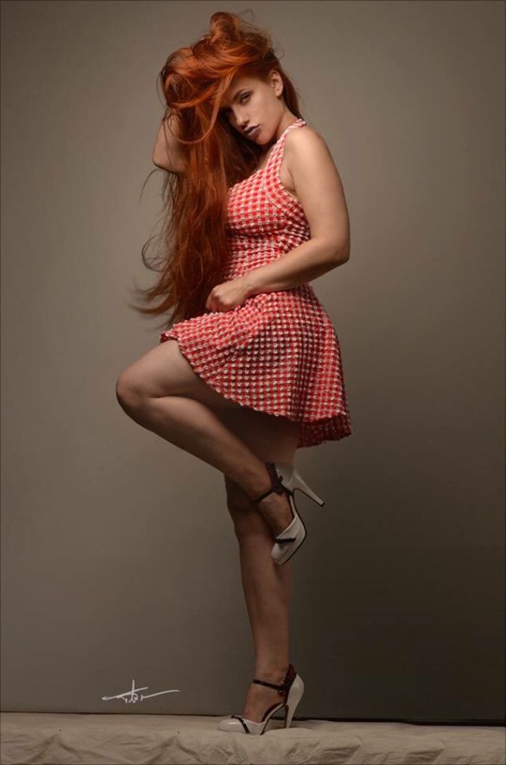 Woman with long red hair in a red gingham dress and high heels posing in a studio