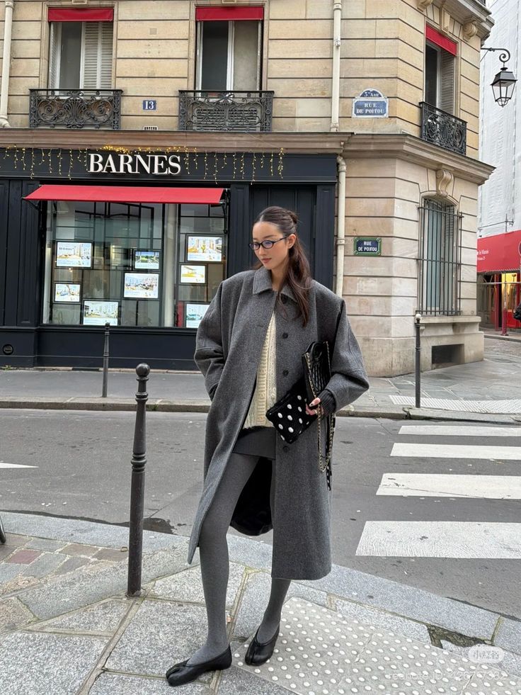 Woman in a long grey coat and glasses standing on a city street