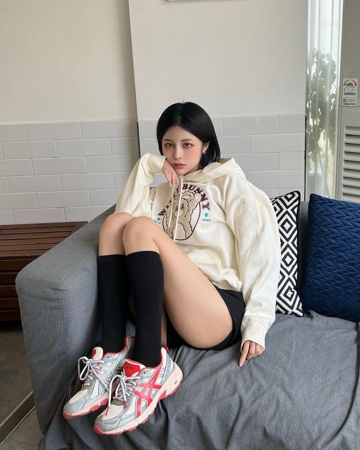 Woman with short black hair wearing a hoodie, shorts, and sneakers sitting on a couch