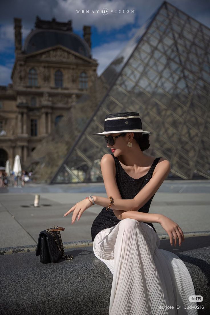 Woman in a black top and white pleated skirt sitting outdoors with a cityscape background