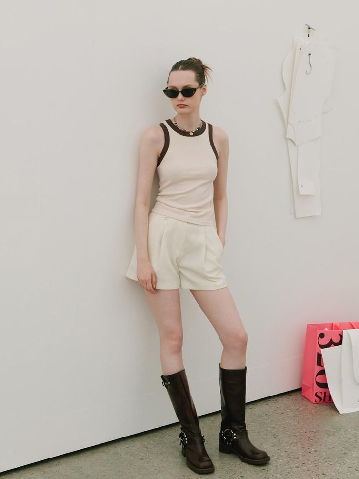 Woman in a tank top, shorts, and knee-high boots leaning against a white wall