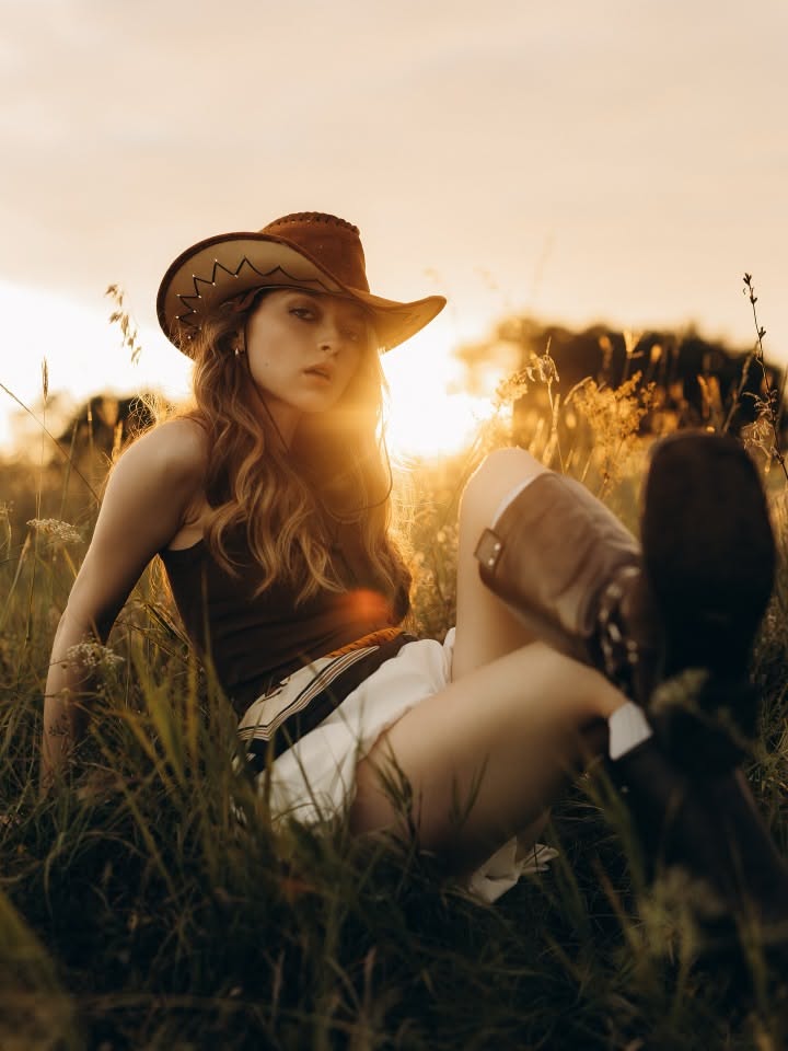Woman in a cowboy hat and boots sitting in a grass field at golden hour