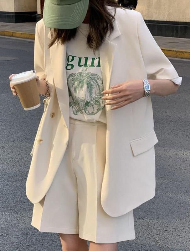 Person in a blazer, shorts, and baseball cap holding a coffee cup outdoors