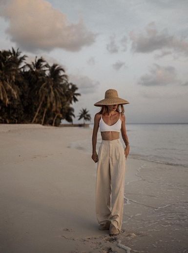 Woman in a straw hat and wide leg pants walking on a beach with palm trees