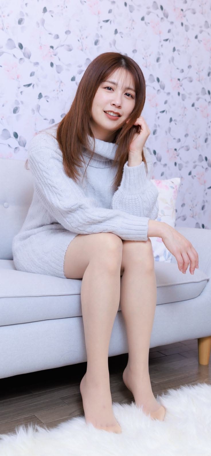 Woman with long hair wearing a light sweater dress, sitting on a sofa indoors