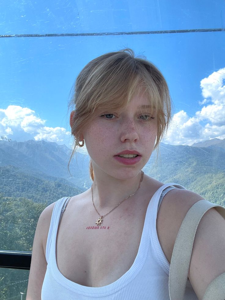 Young woman with blonde hair and freckles in a white tank top outdoors with mountains