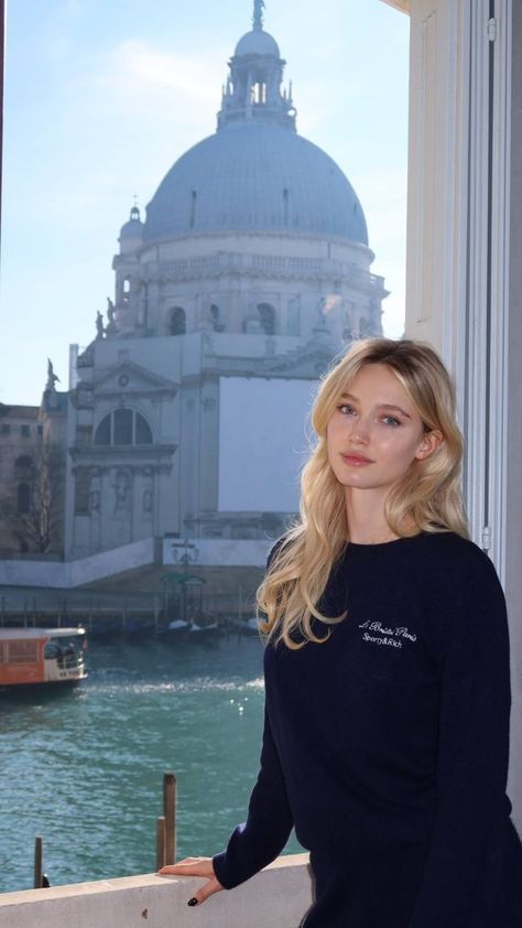 Blonde woman in a navy sweater by a window overlooking a cityscape and water
