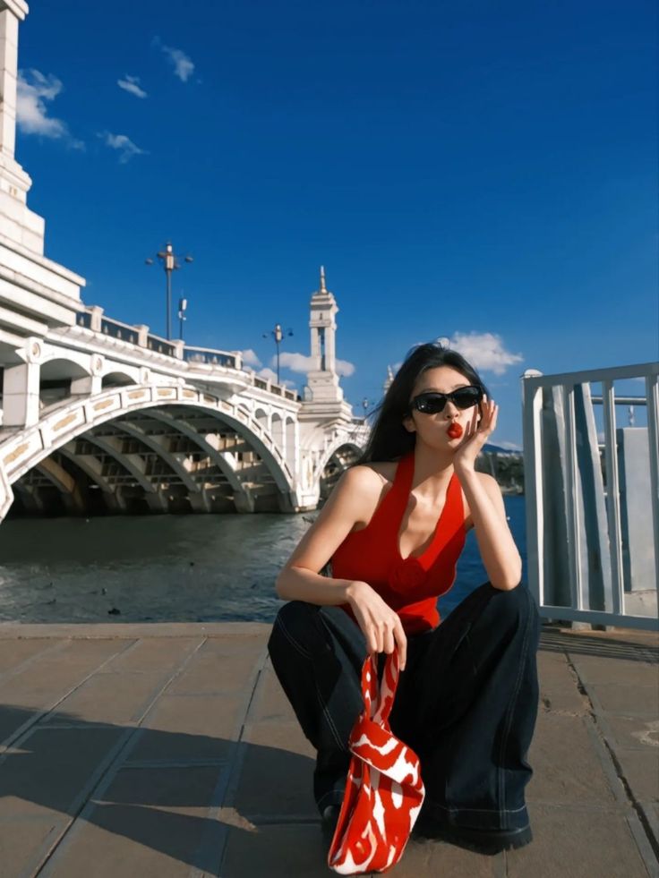 Woman in a red halter top and wide leg pants posing by a river with a bridge
