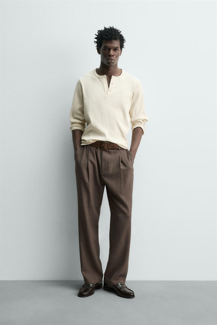 Man in a cream henley shirt and brown pants posing in a studio