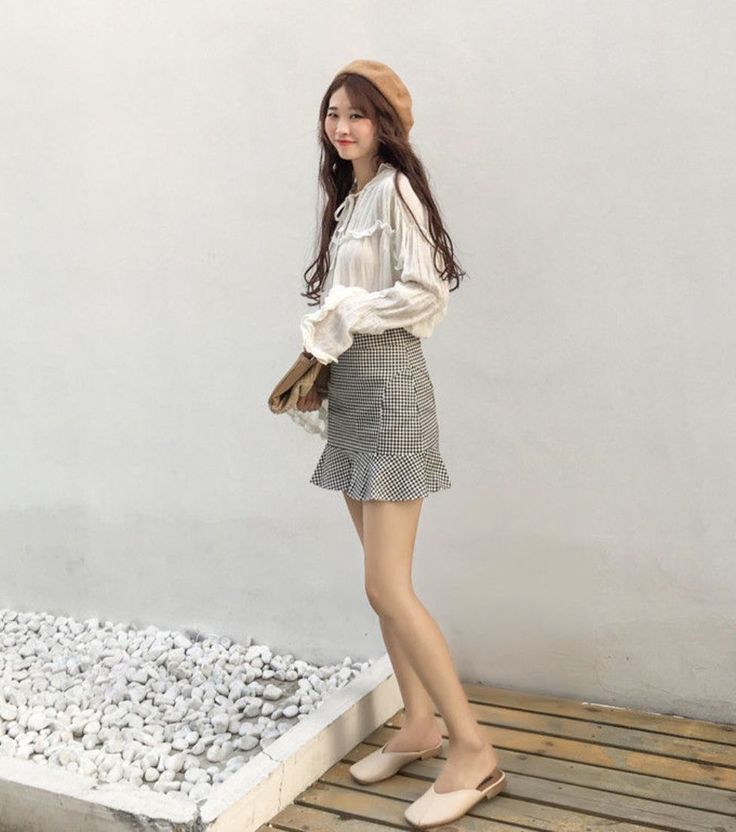 Woman in a beret and gingham skirt standing outdoors against a white wall