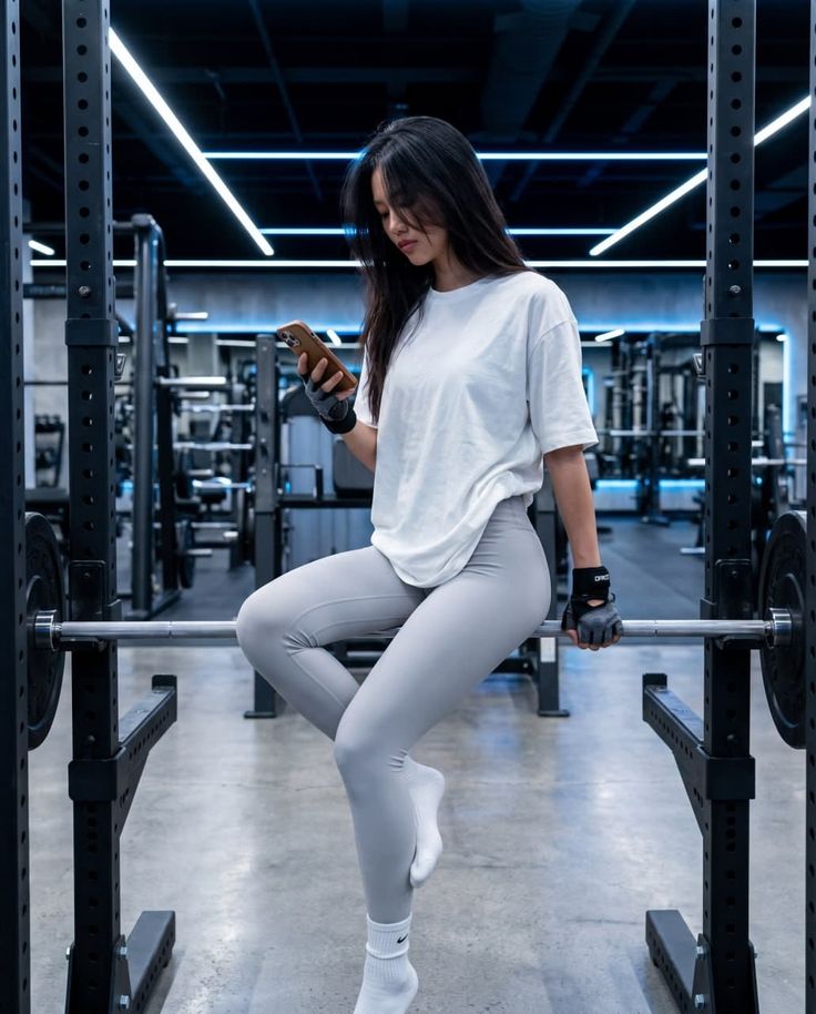 Woman in a gym wearing a white t-shirt and light gray leggings, looking at her phone