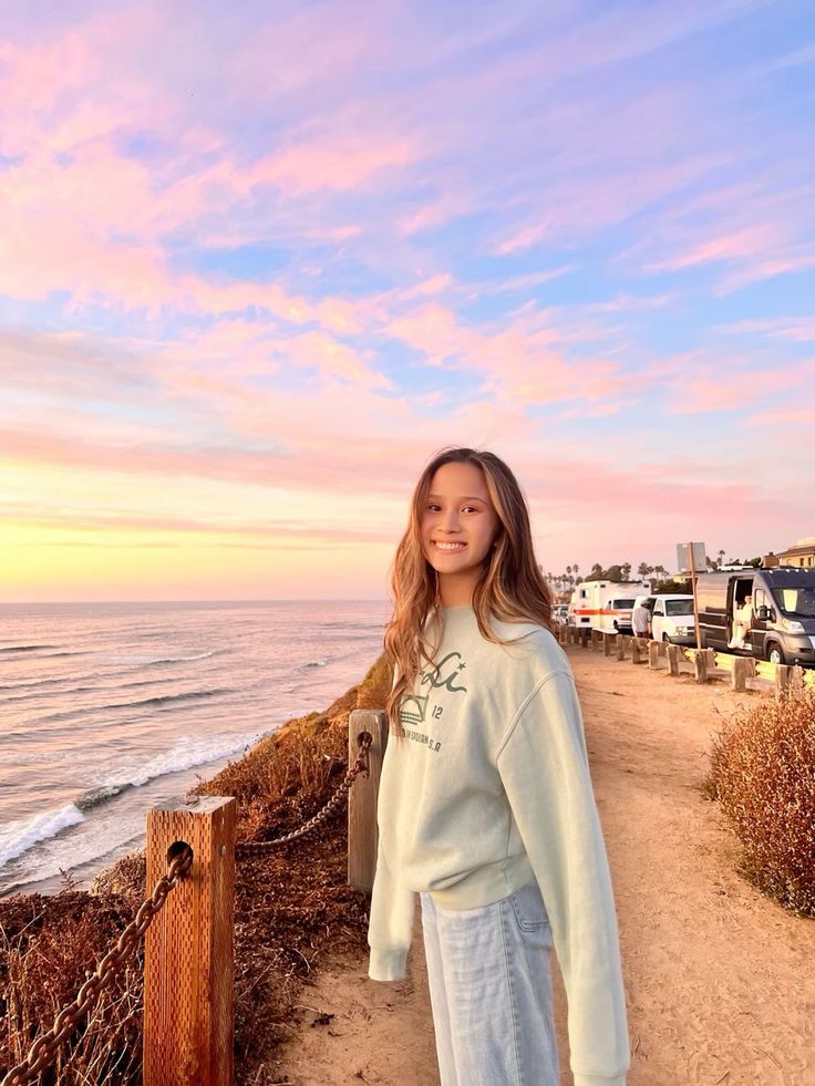 Young woman in a sweatshirt and jeans standing by the ocean at sunset
