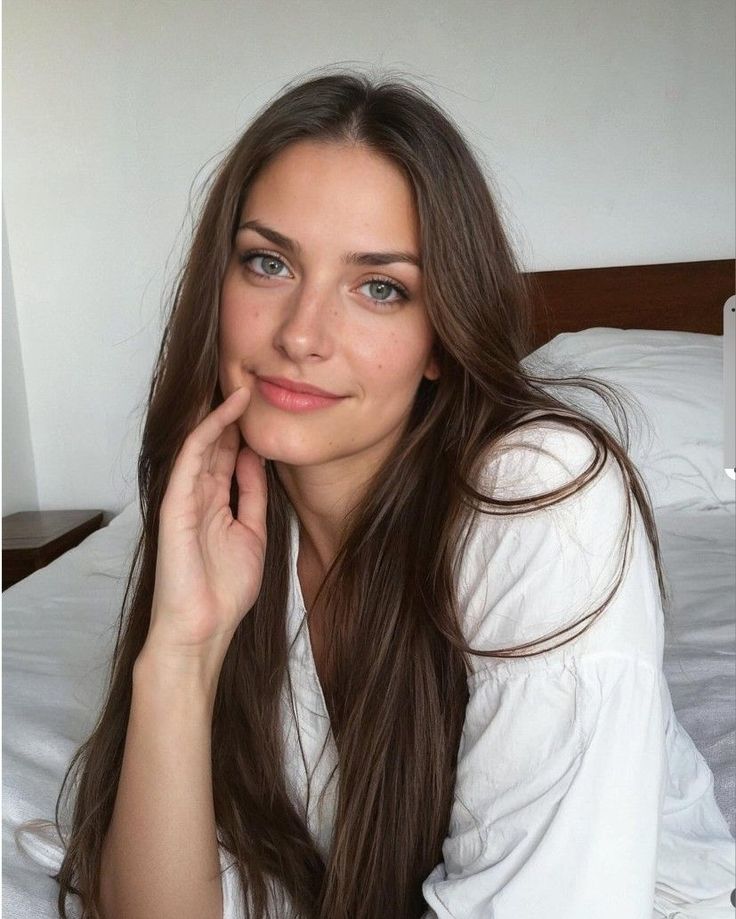 Woman with long brown hair in a white shirt, sitting on a bed