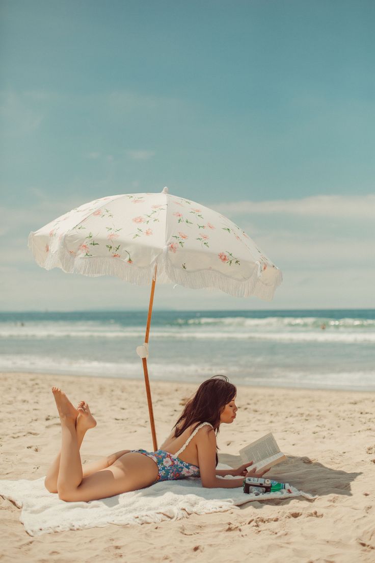 Woman in a floral swimsuit reading a book on a beach under an umbrella