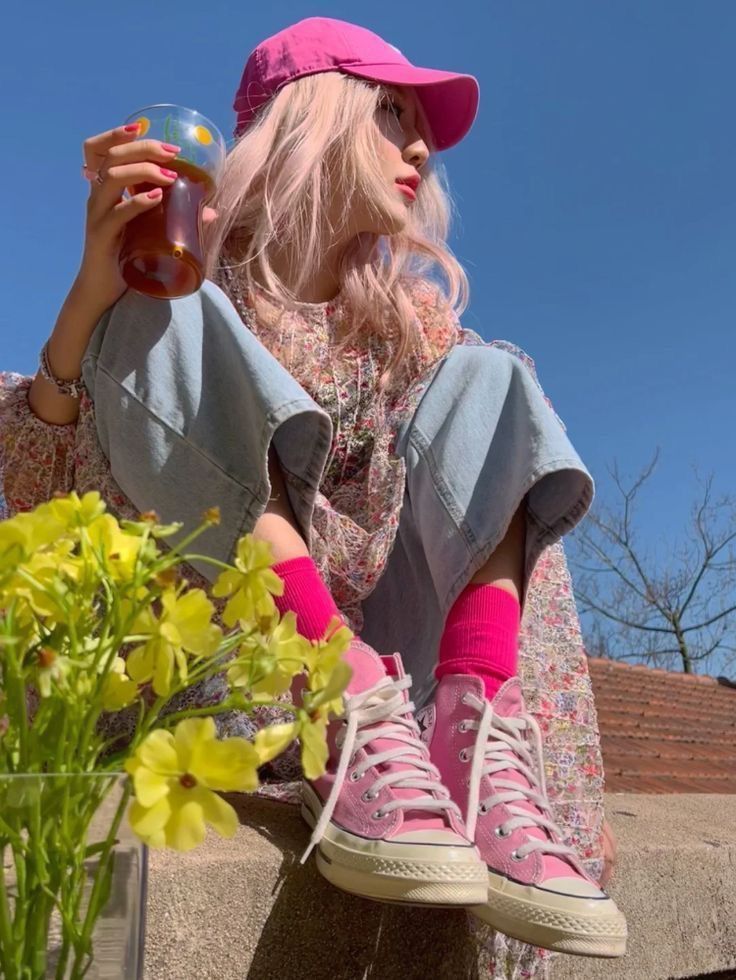Person with pink hair and a pink cap, wearing a floral dress and jeans, holding a drink