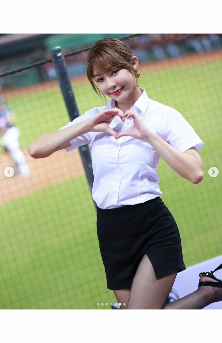 Woman in a white shirt and black skirt making a heart gesture at a baseball field