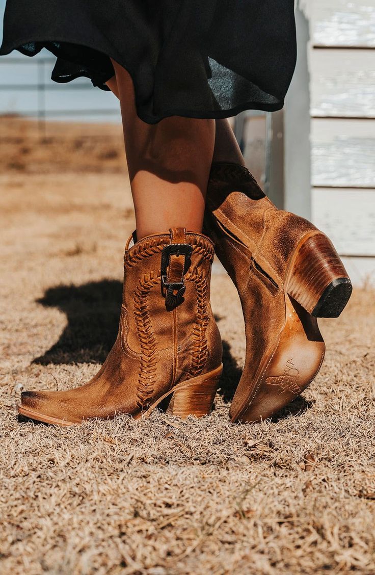 Person wearing brown cowboy boots and a short black dress on dry grass