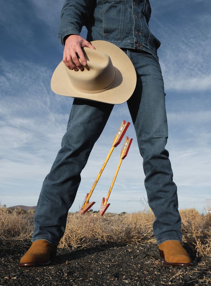 Person in denim holding a cowboy hat, standing over two large arrows in a desert landscape