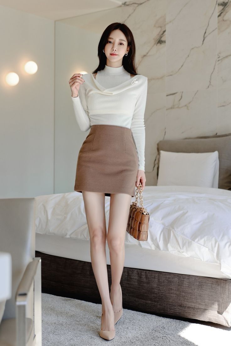 Woman in a white top and brown mini skirt holding a handbag in a bedroom