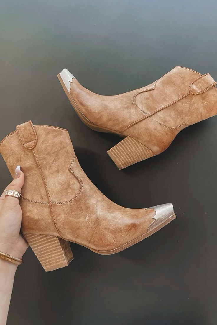 Pair of brown cowboy ankle boots with silver toe caps, held by a hand