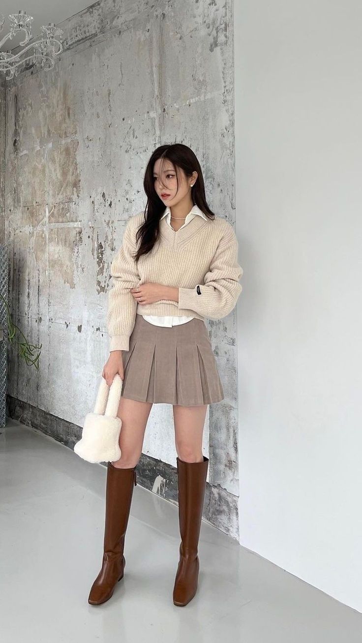Woman in a beige sweater, pleated skirt, and brown boots holding a furry bag