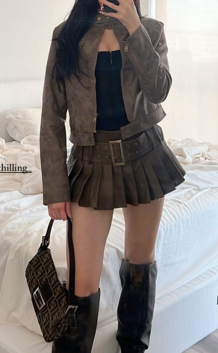 Person in a cropped jacket, pleated skirt, and long boots holding a handbag
