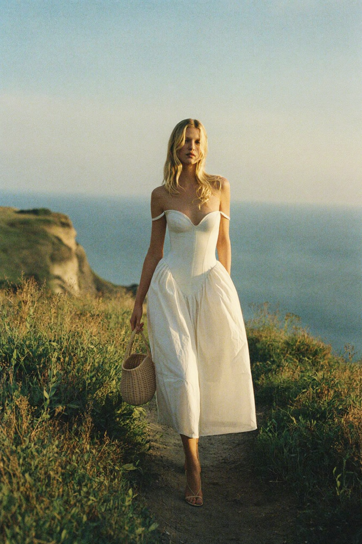 Woman in a white dress walking on a path along a cliffside overlooking the ocean