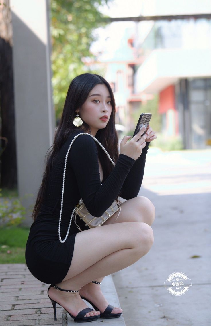 Woman in a black dress and high heels squatting outdoors while holding a phone