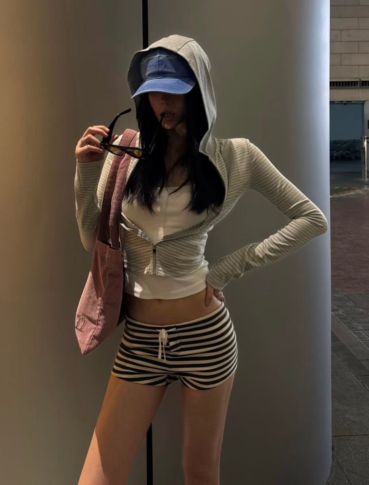 Woman in a hoodie, baseball cap, and striped shorts posing outdoors