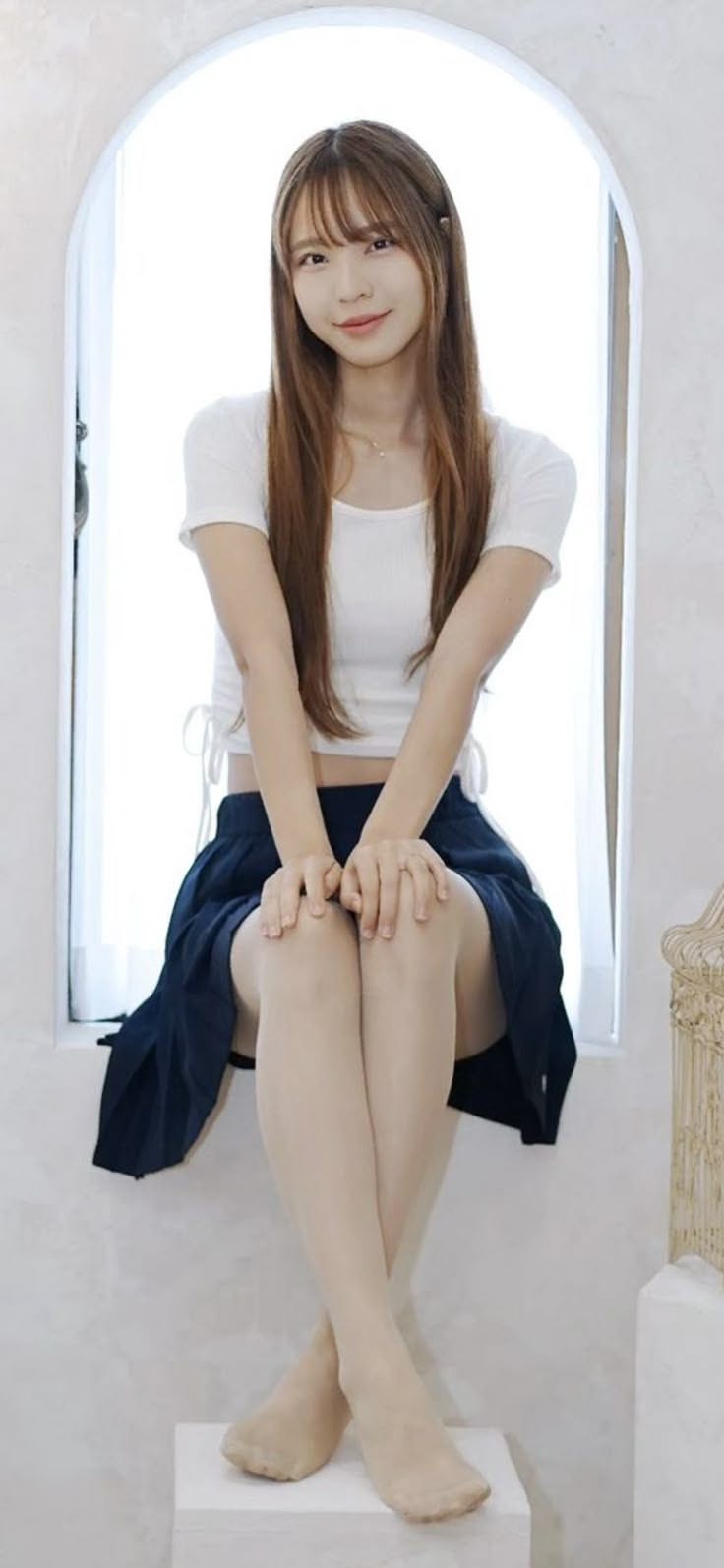 Woman with long hair in a white crop top and navy skirt sitting by a window