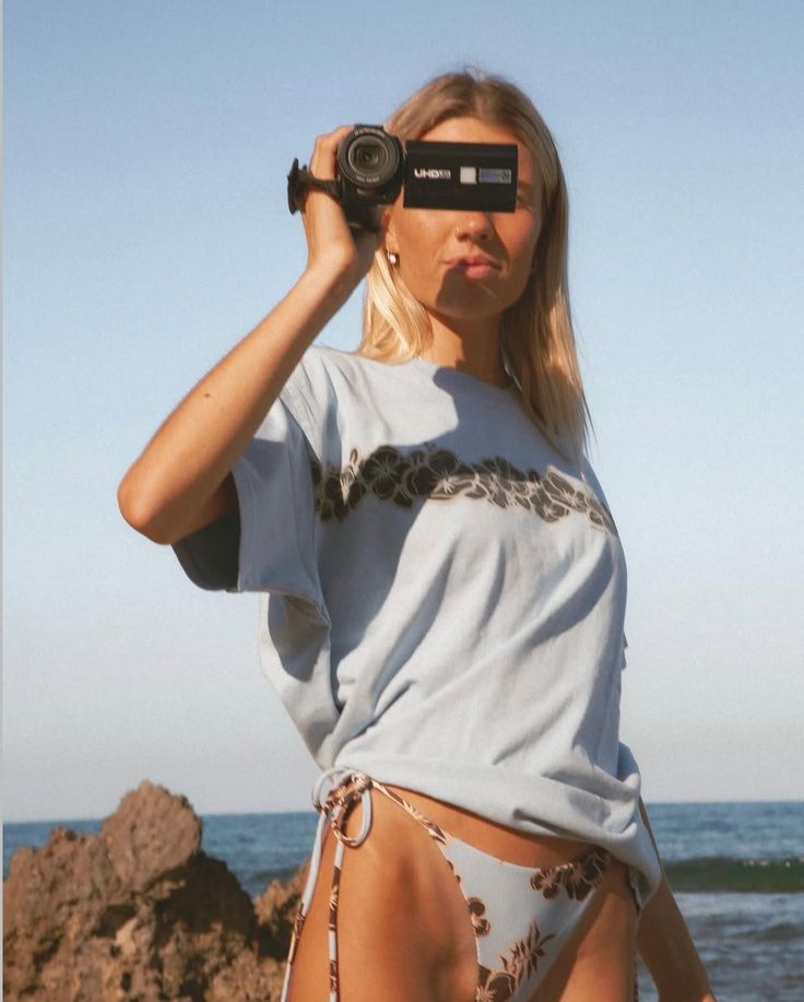Blonde woman in a bikini and oversized t-shirt holding a camera at the beach
