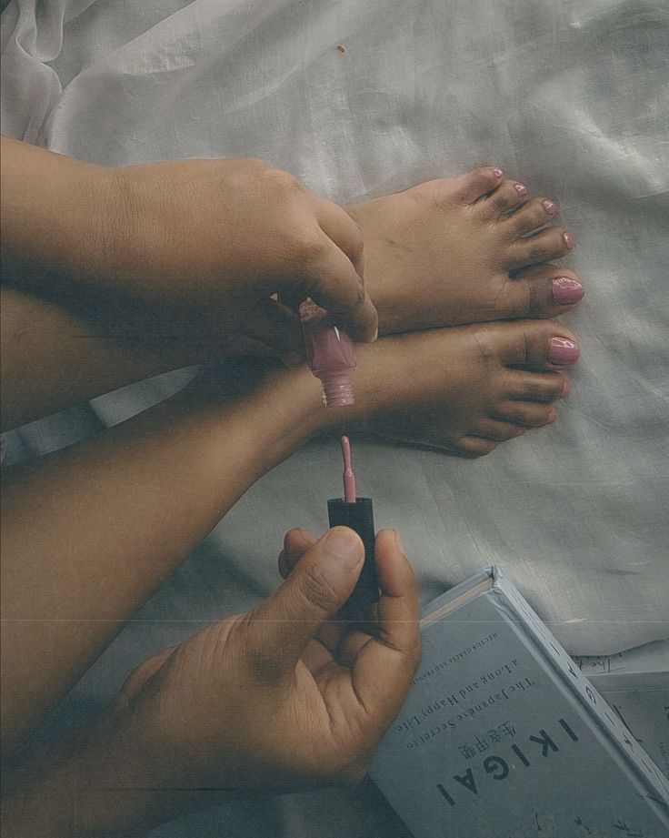 Person painting their toenails pink while holding a book
