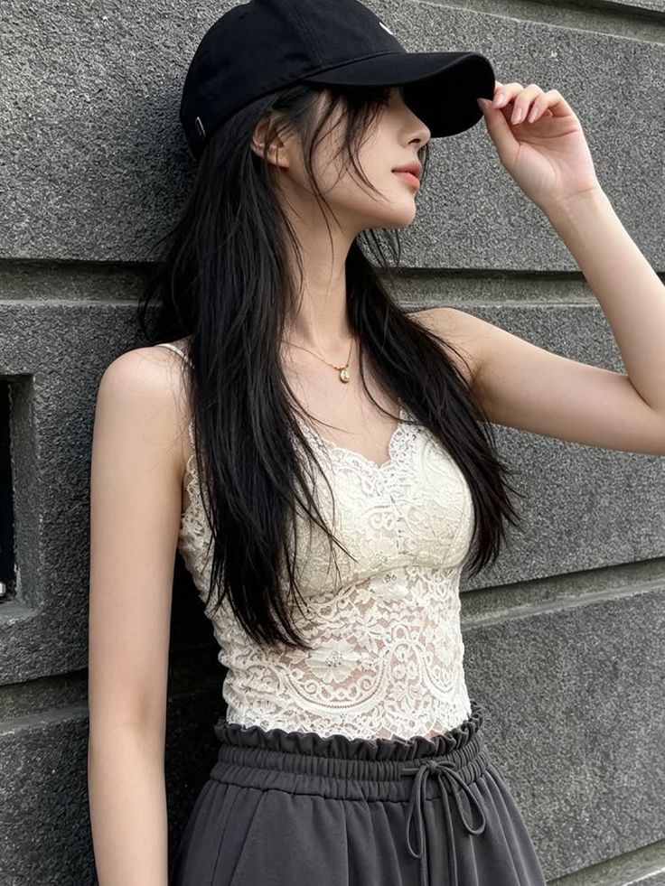 Woman in a black cap and lace top leaning against a gray wall