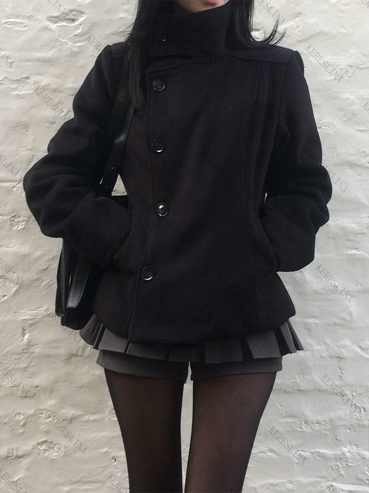 Person in a black coat, pleated skirt, and tights against a white brick wall