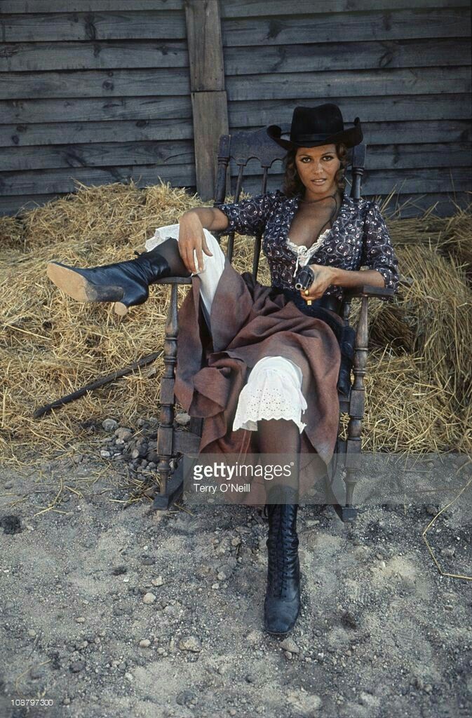 Woman in a cowgirl outfit and hat sitting in a chair with a gun